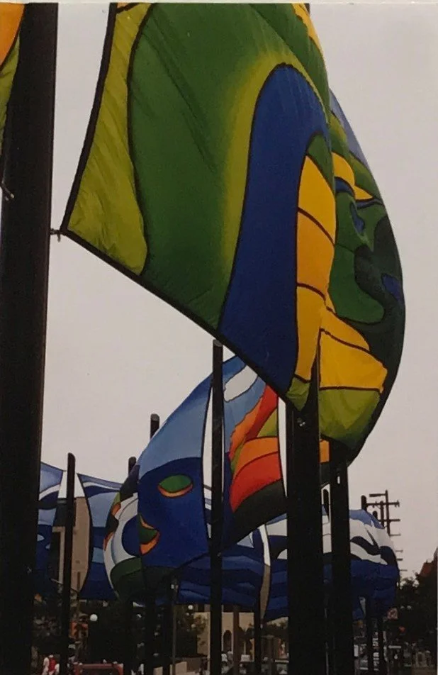 Multiple colorful flags fluttering on tall black poles in an outdoor setting. NCC banner competition winner Jane Thompson, 1988 Ottawa Ontario Canada.