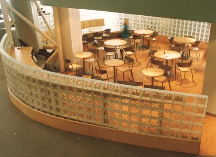 Indoor dining area with multiple round wooden tables and chairs, separated by glass block walls, with a staircase nearby.