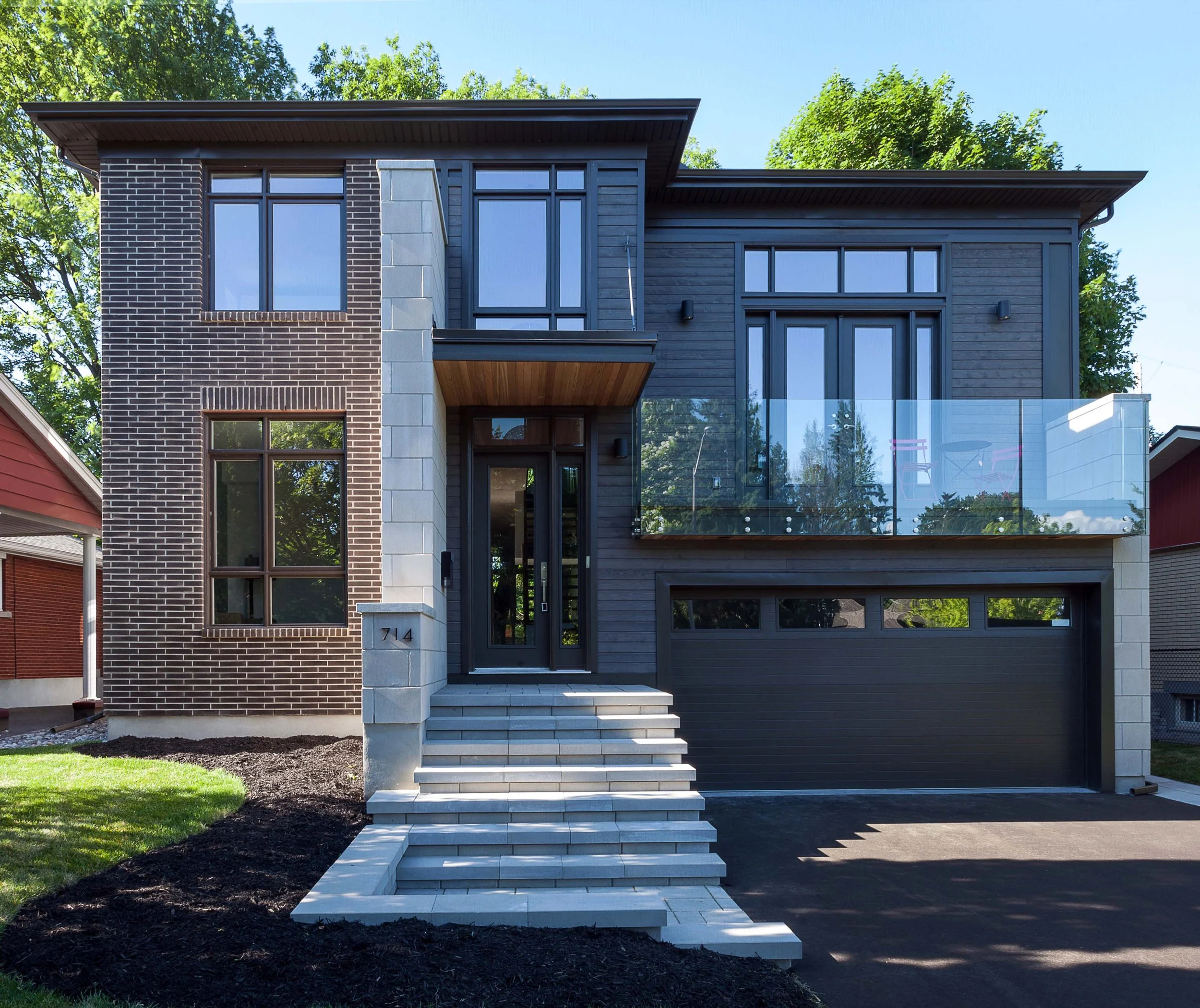 Front view of a modern, two-story house with dark wood and brick exterior, large windows, glass balcony, and a black garage door, with a staircase leading to the front door.