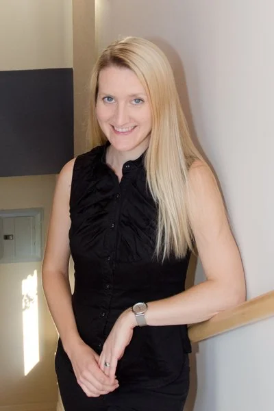 A smiling woman with long blonde hair wearing a sleeveless black blouse and a watch, leaning against a staircase railing indoors.
