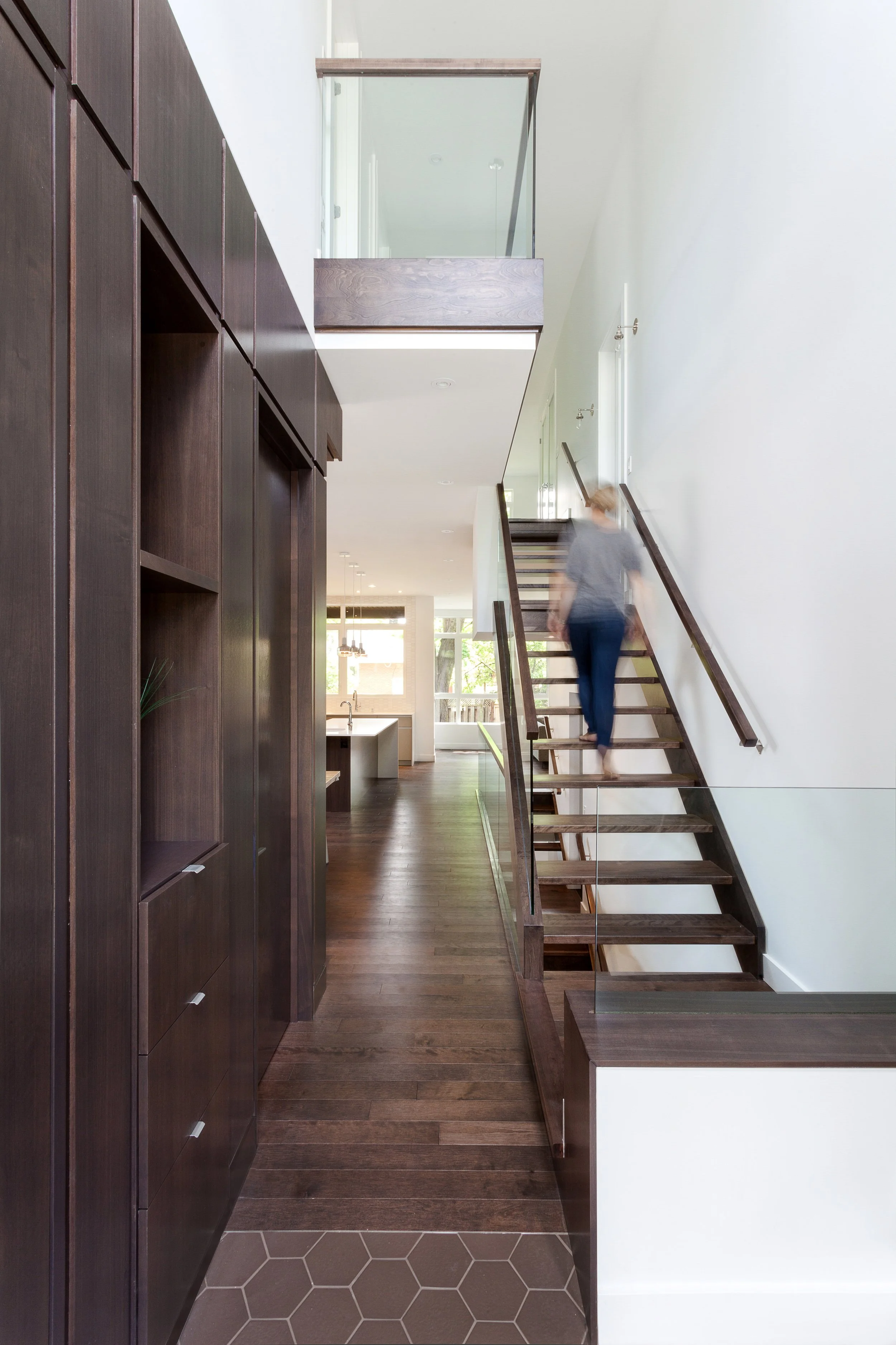 Interior view of a modern home with dark wood flooring, a staircase with wooden steps and glass railing, and white walls. A blurred woman is walking up the stairs near a kitchen area with large windows in the background.