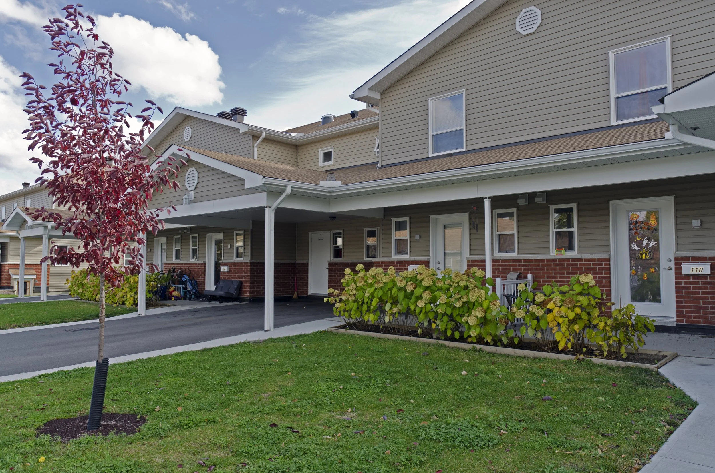 Multi-family residential buildings with beige siding and red brick, a small tree with reddish leaves in the front yard, green shrubbery, and a walkway.