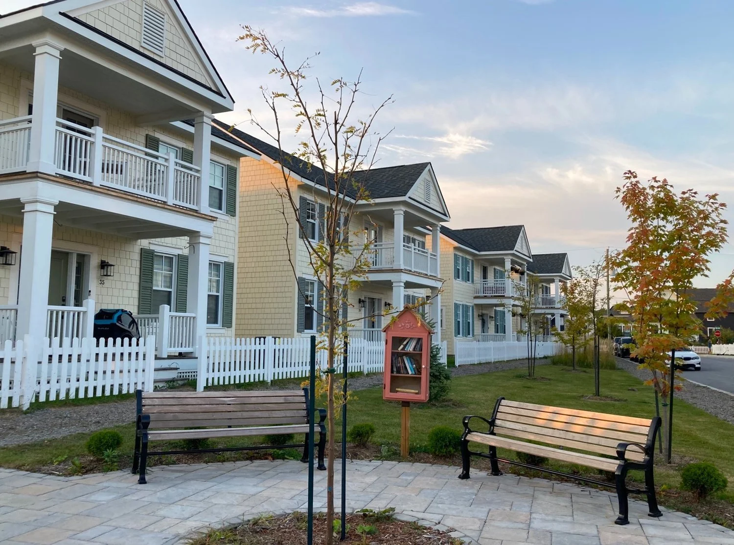 A peaceful residential neighborhood with new multi-story houses featuring balconies, white fences, and a small park area with benches, young trees, a small pink book-shaped library box, and a paved walkway under a clear sky.
