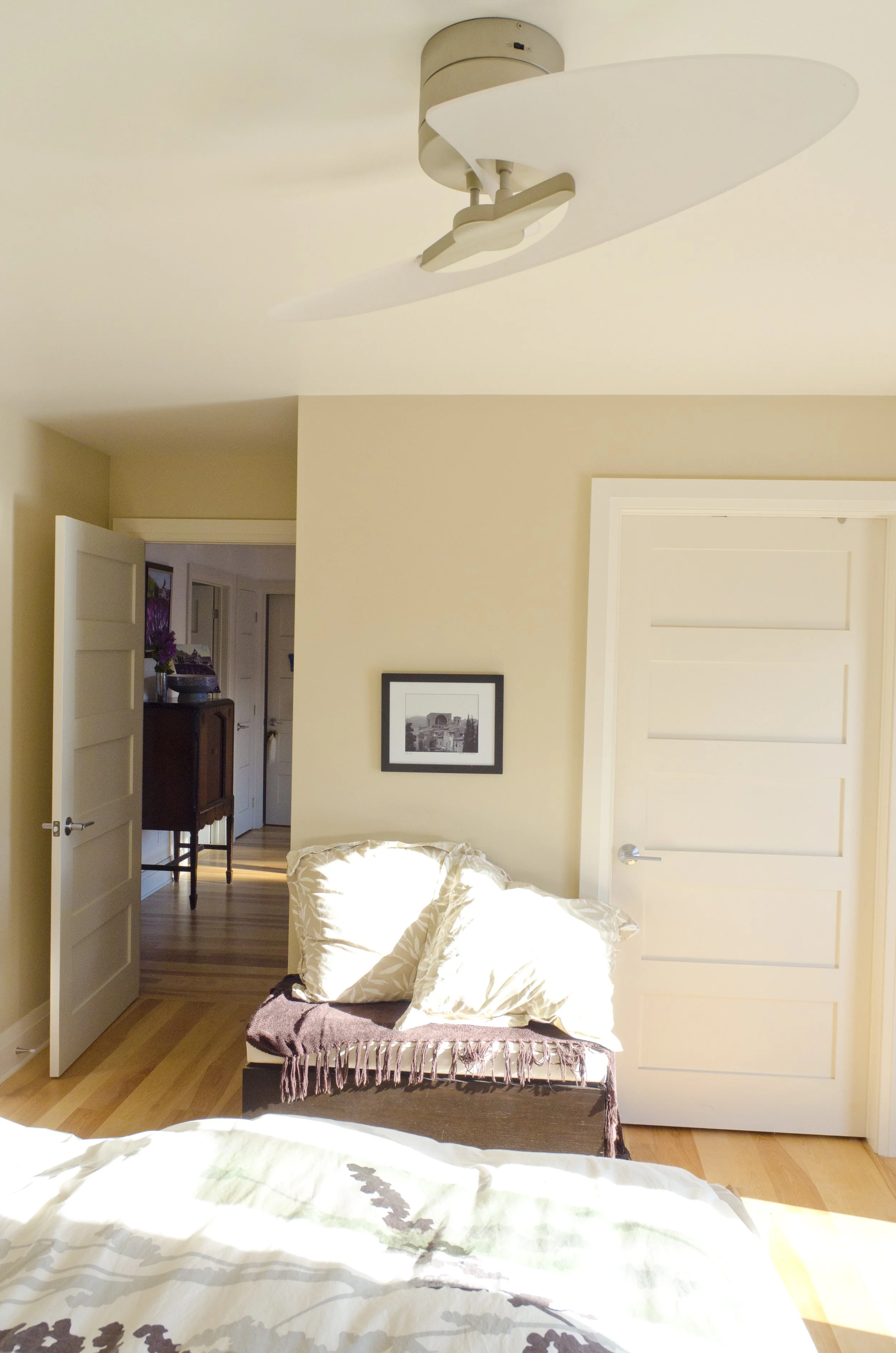 Sunlit bedroom with ceiling fan, framed black-and-white photo, cream pillows, wooden floor, open door leading to hallway with furniture.