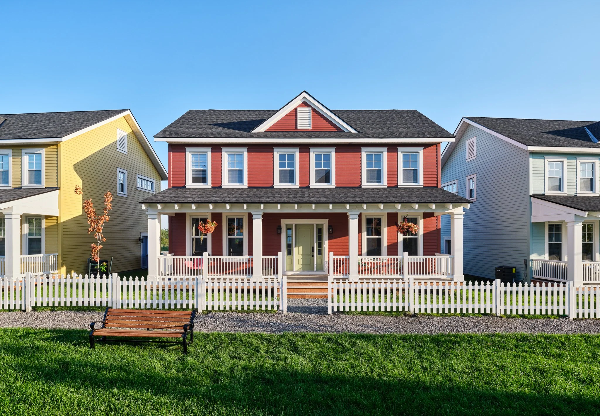 Front view of a red, two-story house with a porch and white railings, flanked by a yellow house on the left and a light blue house on the right, with a green lawn and a wooden bench in the foreground, and a clear blue sky above.