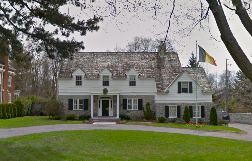 A large, historic house with a stone and white wood exterior, multiple windows with black shutters, and a steep shingled roof. A flagpole with a flag stands in front of the house, surrounded by a well-maintained lawn and garden.