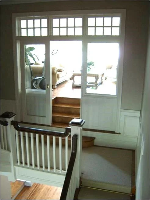 View of a staircase leading to a living room with couches, chairs, and a coffee table illuminated by sunlight through large windows.