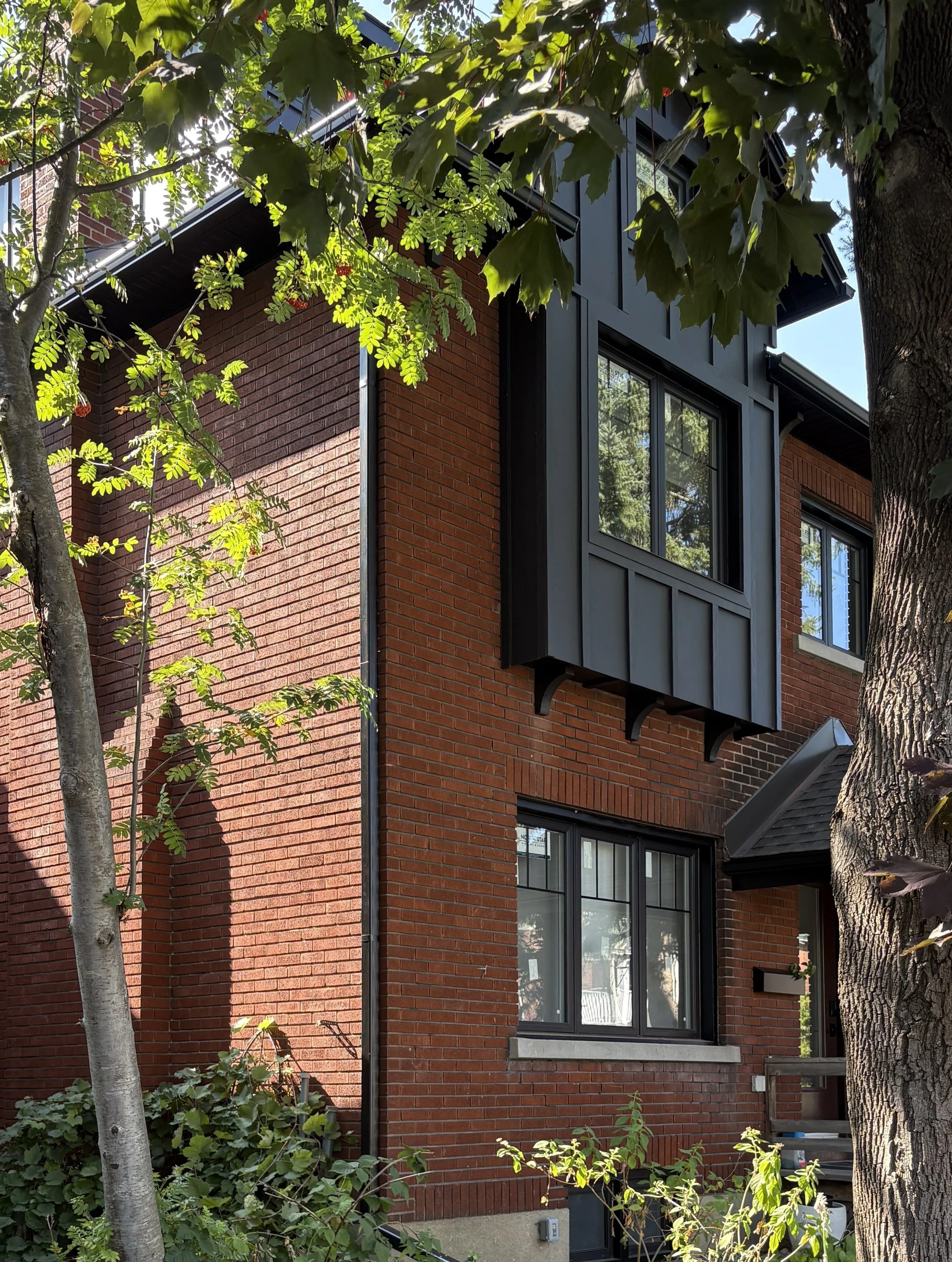 Part of a red brick house with black window frames, trees with green leaves, and sunlight filtering through the leaves.