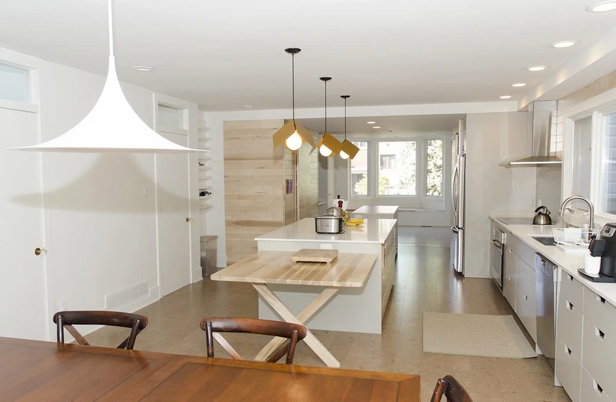 Modern kitchen with white walls, wooden flooring, and large windows. Contains a kitchen island with a white countertop and pendant lights, along with stainless steel appliances and a dining table with chairs.