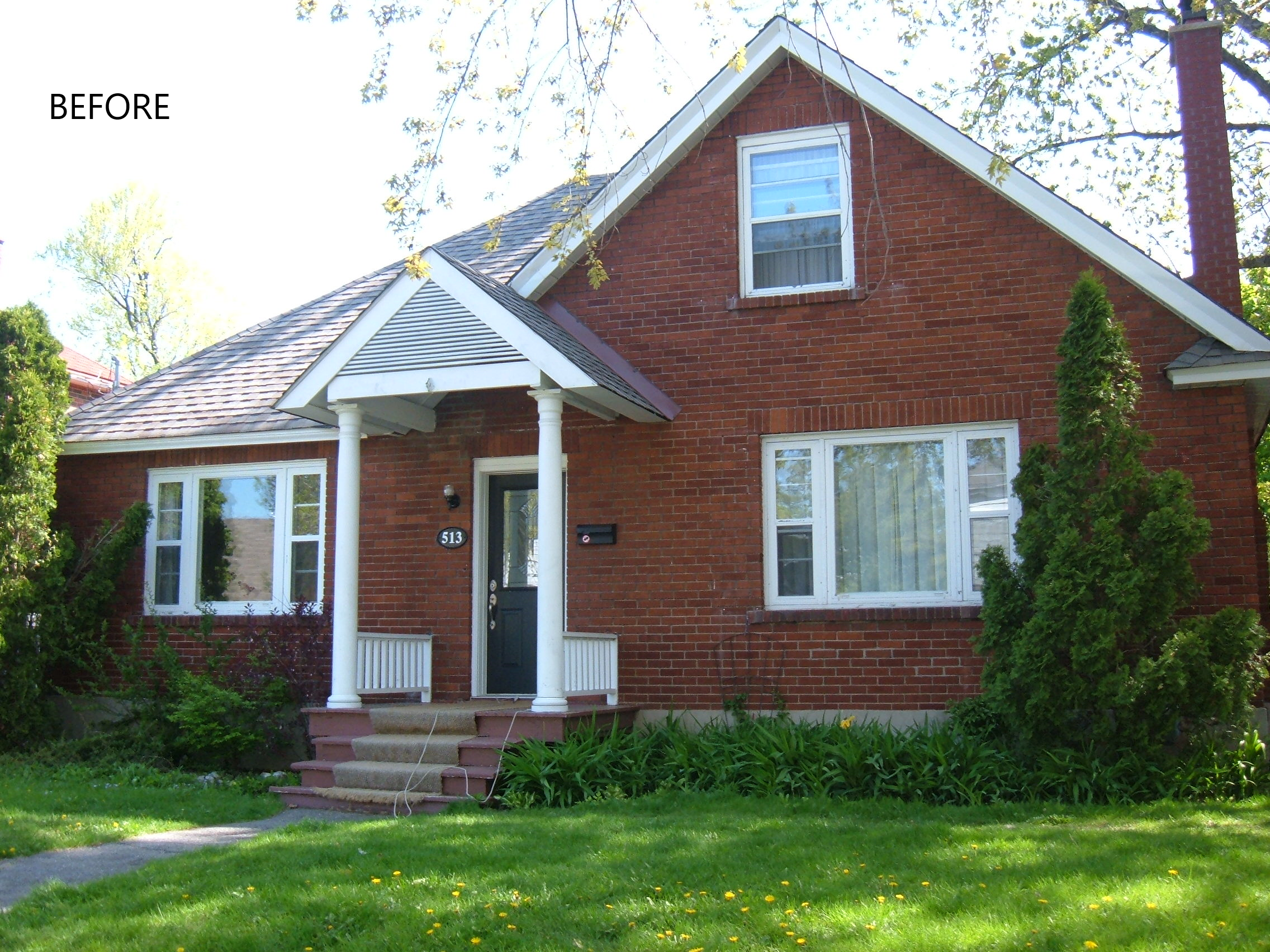 A brick house with white trim, a front porch with four steps, two white columns, and a small black door. There is a large window to the left of the porch and a smaller window above the door. The house has a gabled roof with grey shingles, and there are bushes and a green lawn in front of it.