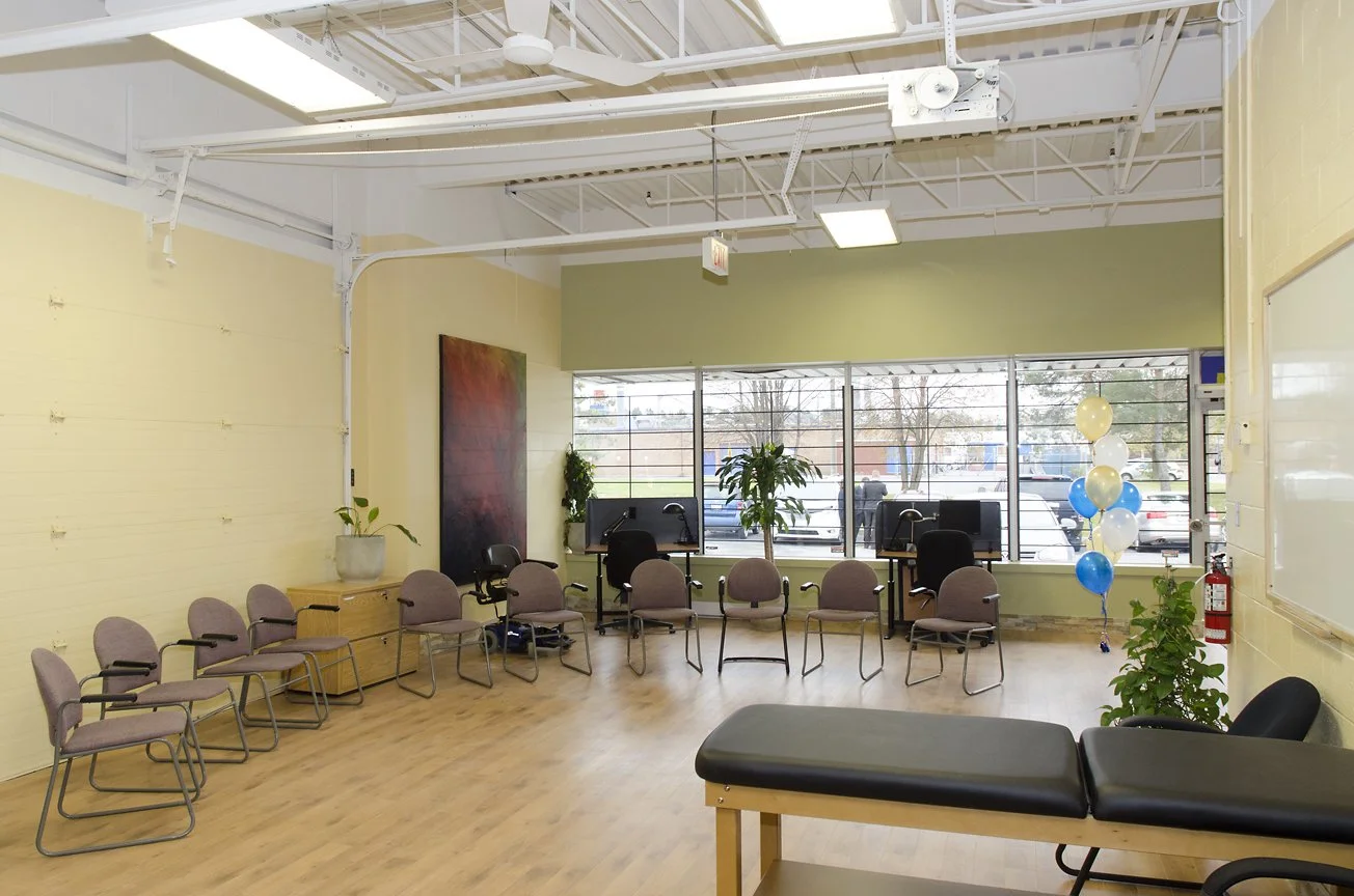 A waiting room with a row of chairs along the left wall, some chairs around two computer desks near the window, and a black examination table on the right side. There are balloons and potted plants, and large windows letting in natural light.