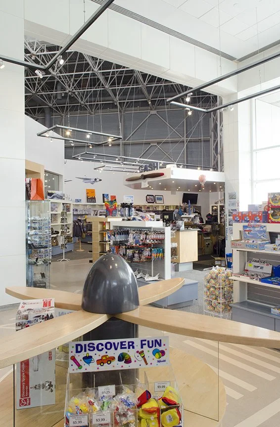 Indoor toy store with a large model of an airplane on a display table in the foreground, shelves filled with toys and games, and a high ceiling with exposed metal beams.