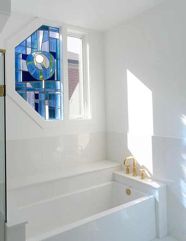 Bathroom with a white bathtub, gold fixtures, a stained glass window with a circular design, and natural sunlight streaming in.