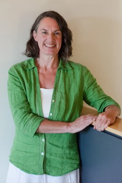 A smiling woman with shoulder-length dark hair wearing a green button-up shirt over a white top, standing indoors against a beige wall, resting her hands on a counter.