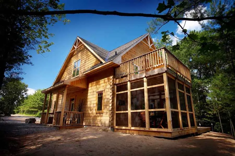A newly constructed energy star log home in a forested area with a clear blue sky overhead. The house features a high-pitched roof, a front porch, and a large screened-in porch or sunroom on the side.