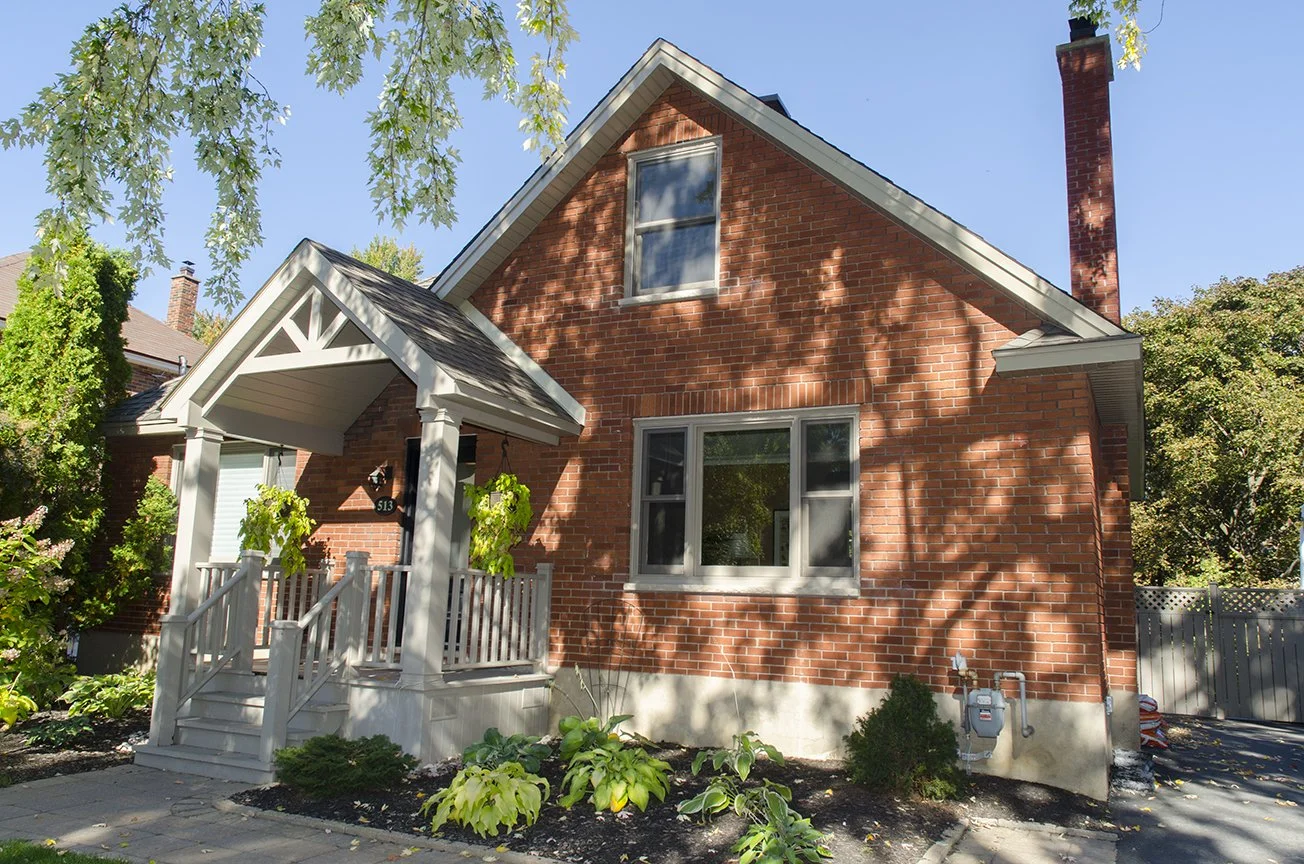 A two-story brick house with a front porch, surrounded by trees and garden plants, with shadows of leaves cast on the facade.