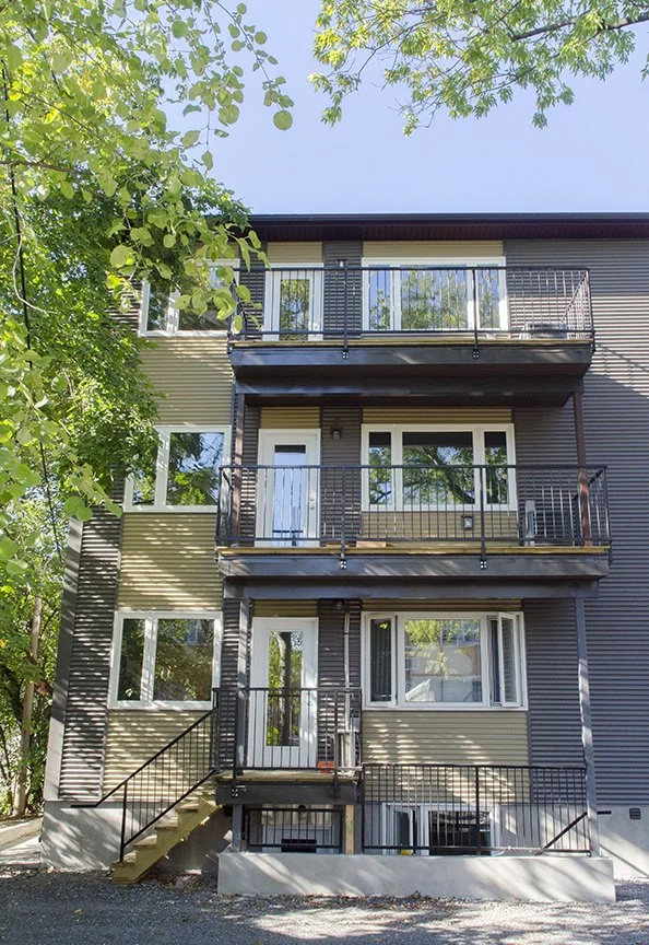 Three-story residential building with balconies, beige and dark gray exterior, surrounded by green trees.