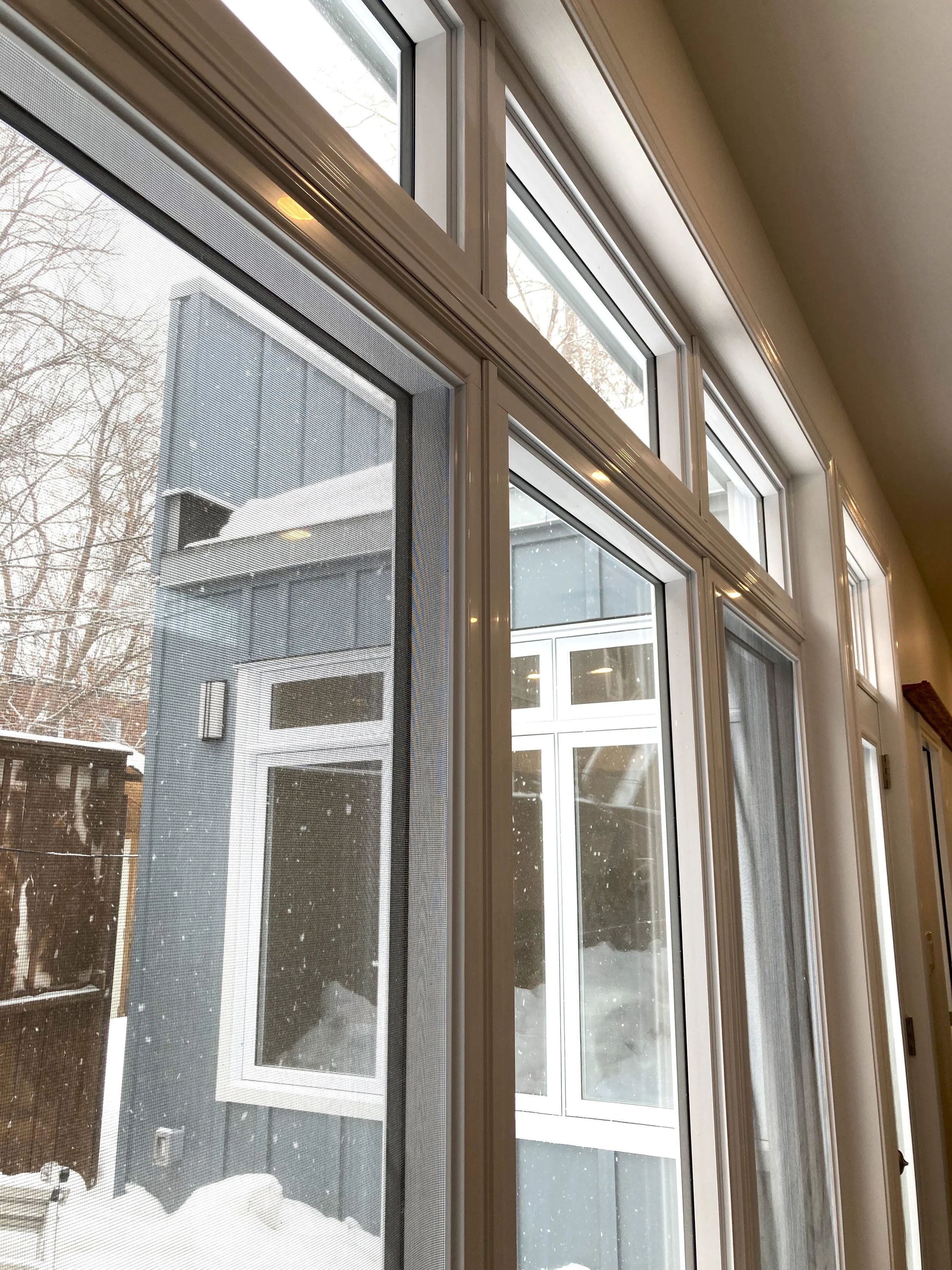 Interior view of large window with multiple panes looking out onto a snow-covered backyard with a neighboring building and a wooden fence.