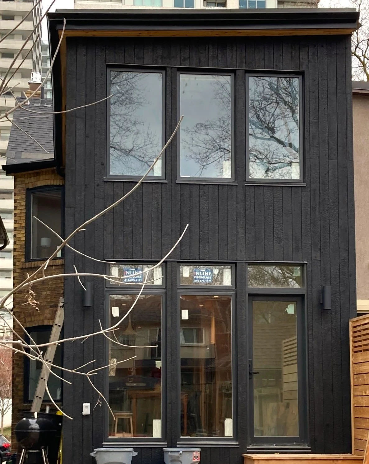 Front view of a three-story modern black wooden house with large windows, some of which have signs that say 'LINE FIBERGLASS'. There is a small tree with bare branches in the foreground and a ladder leaning against the house on the left side.