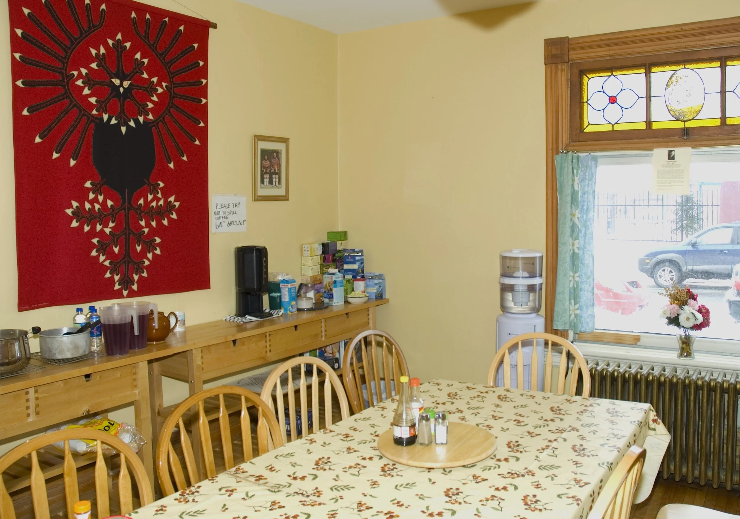 A cozy dining area with a wooden table covered in a floral tablecloth, surrounded by wooden chairs. On the table are condiments and bottles. To the right, there's a window with a floral curtain, a vase of flowers, and a radiator beneath. A water dispenser is near the window. On the left wall, there's a large red tapestry with a black design, a small framed picture, and a sign that says, "Please try not to spill coffee or damage." A sideboard holds supplies and drink stations.