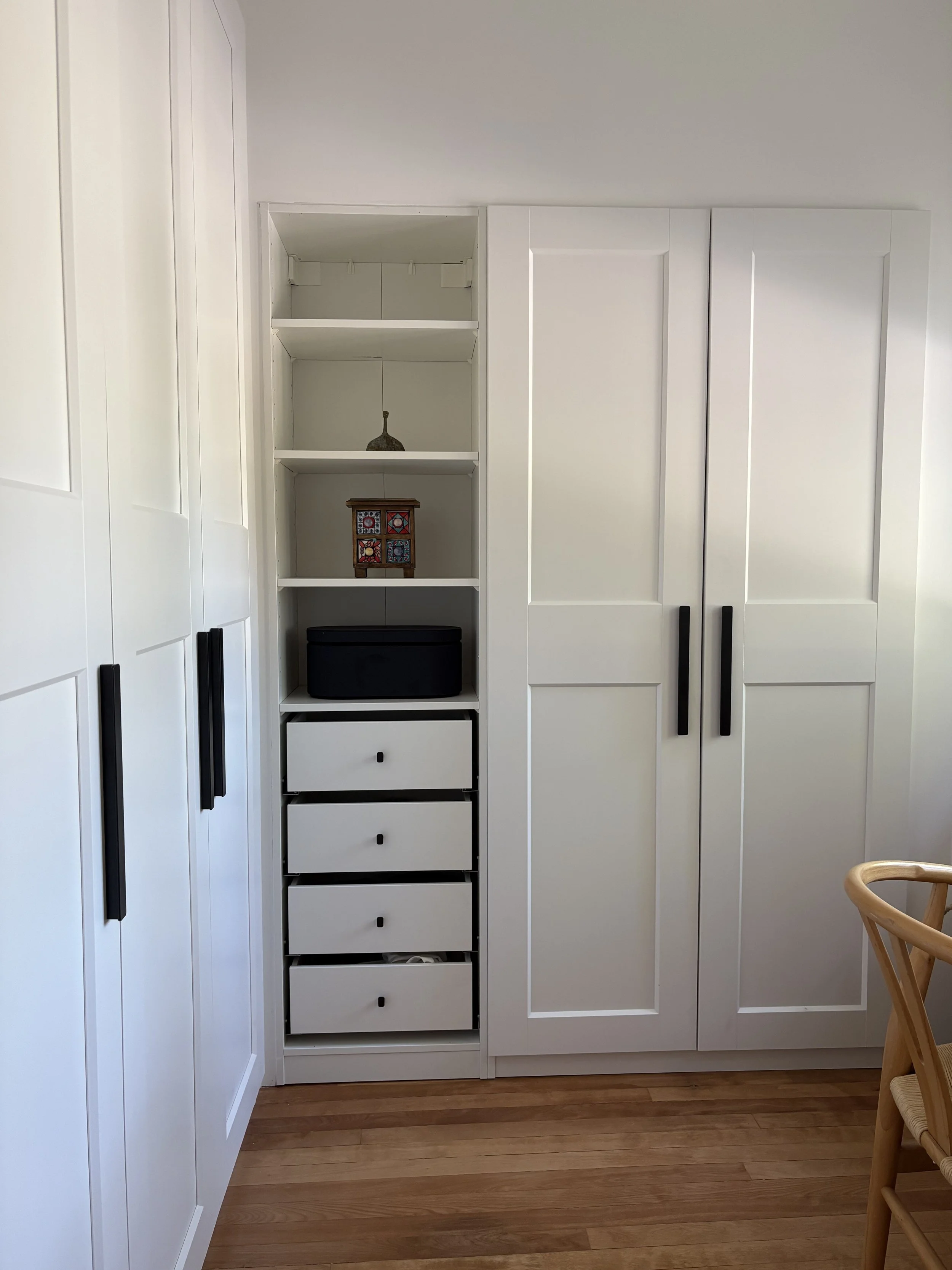 A white built-in closet with black handles, featuring multiple shelves, drawers, and cabinets, in a room with wooden flooring and a partial view of a chair on the right.