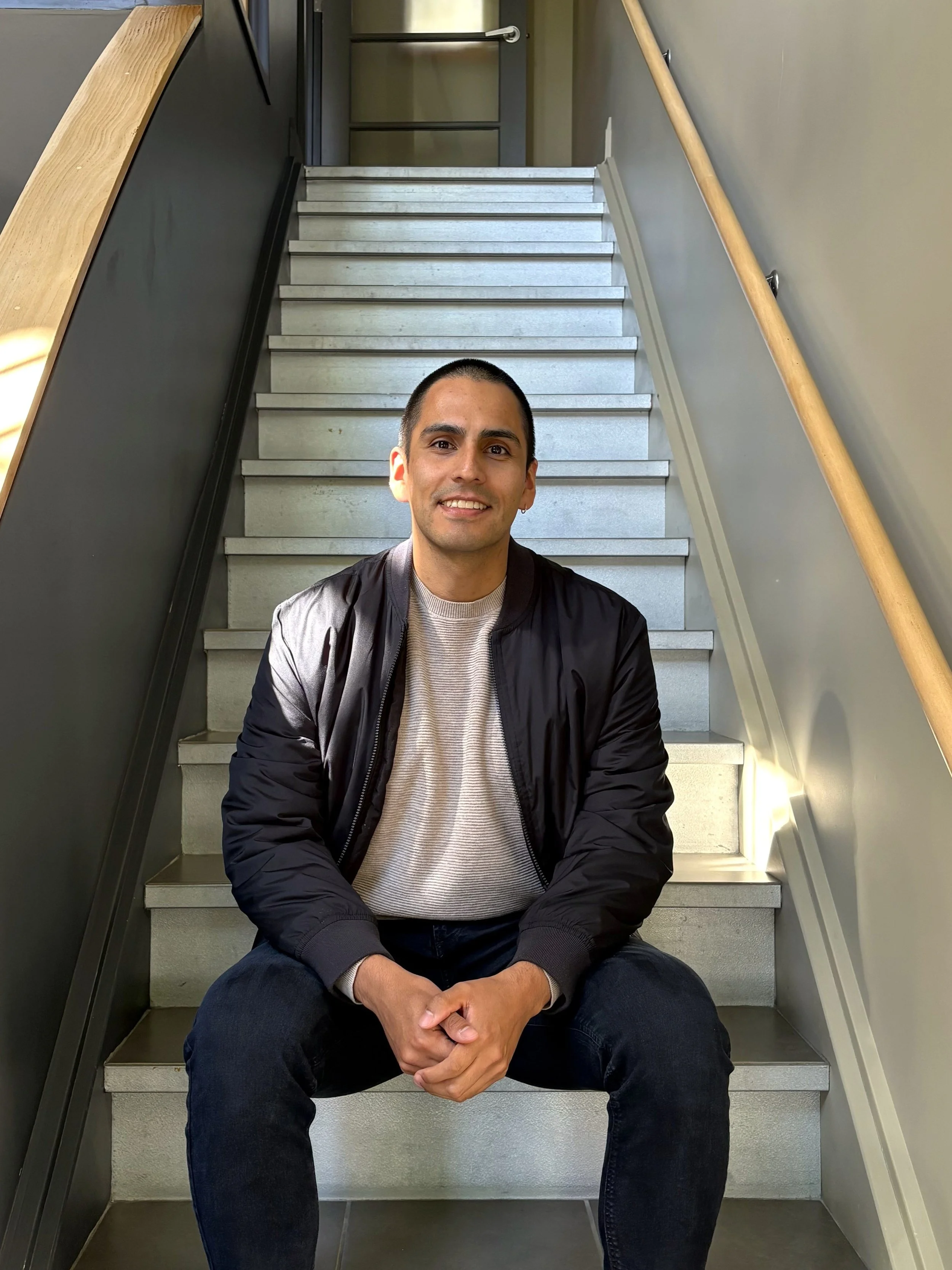 A young man with short dark hair sitting on a staircase, smiling at the camera, wearing a black jacket, light-colored shirt, and dark jeans.