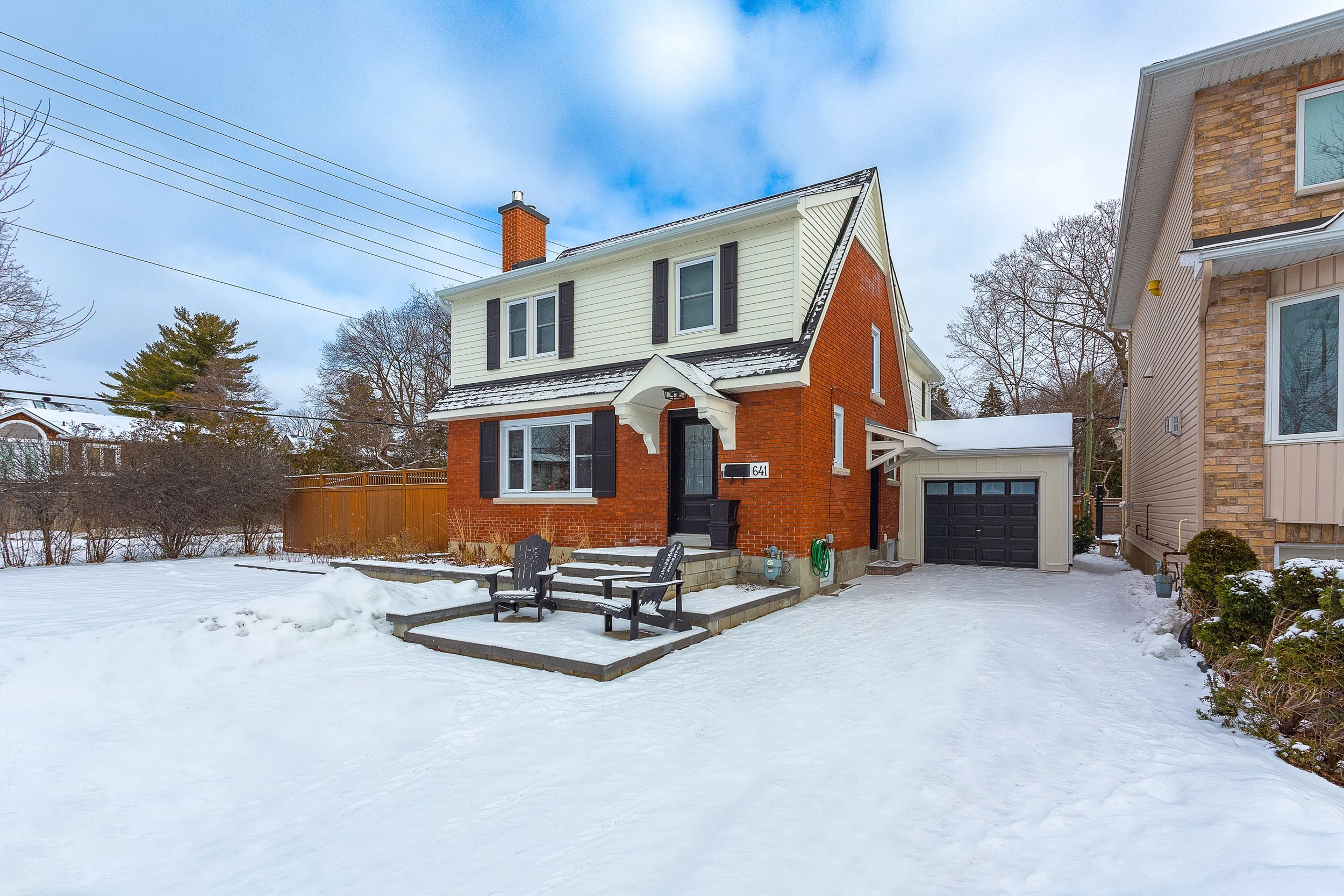 A brick house with a white upper floor and black shutters on the windows, set in a snowy landscape with outdoor seating and neighboring houses visible.
