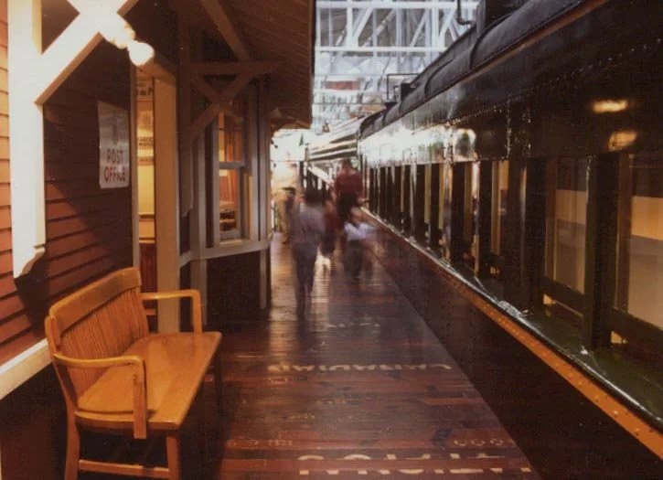 People walking on a train station platform at sunset, with a vintage train on the right side and a wooden bench on the left.