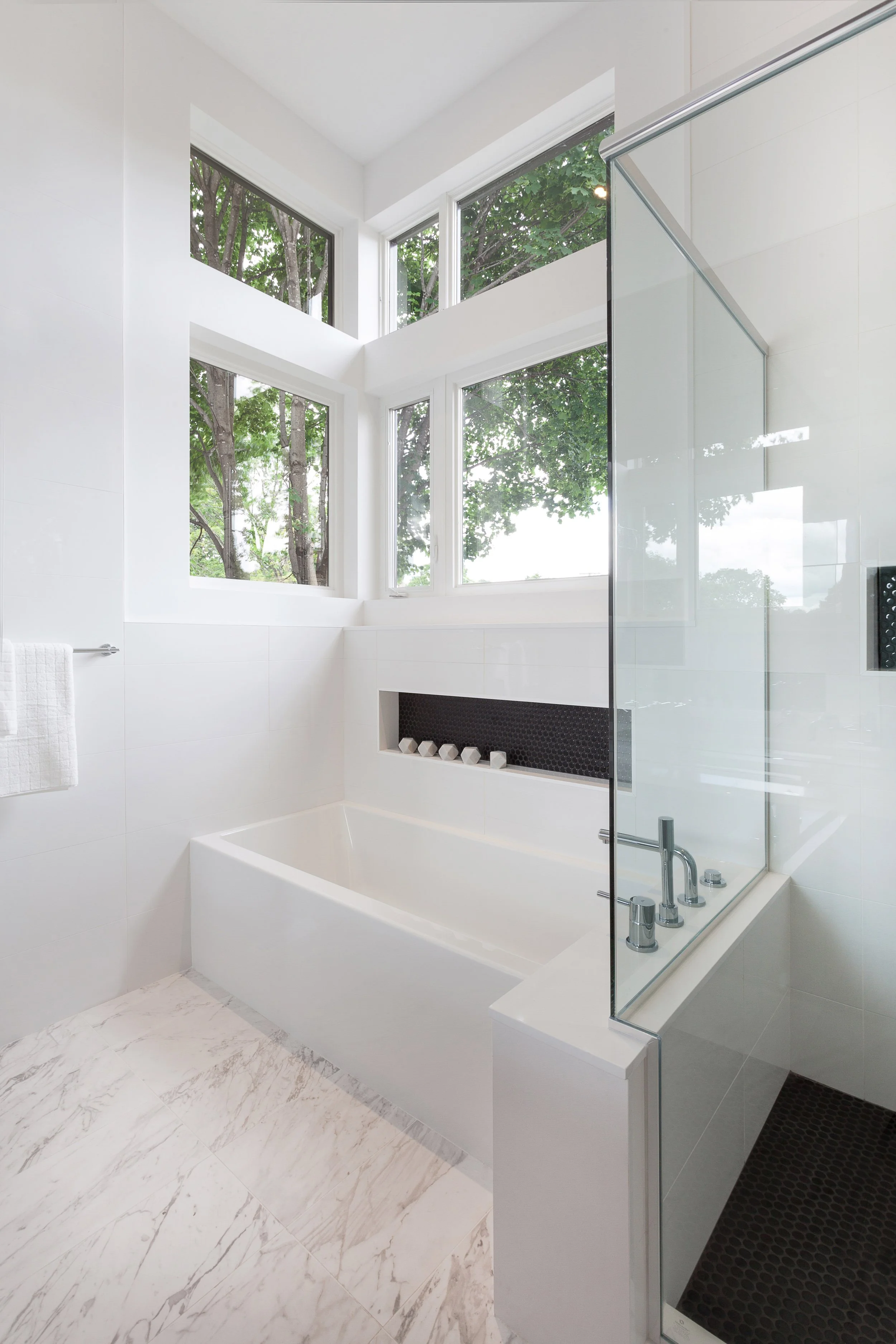 Bright bathroom featuring a white bathtub, large corner windows showing outdoor trees, and a glass shower enclosure.