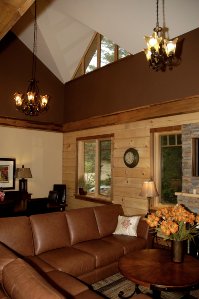 Living room with exposed log exterior walls, a brown leather sofa, a wooden coffee table with a vase of orange flowers, windows showing greenery outside, and a fireplace.