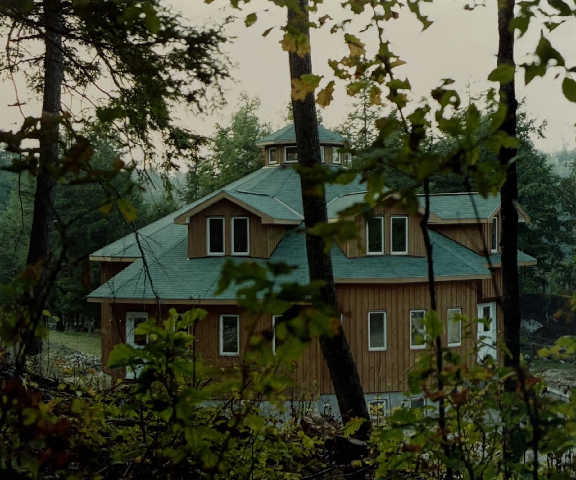 A wooden retreat house with a green metal roof, surrounded by trees with leaves, seen through branches and foliage.