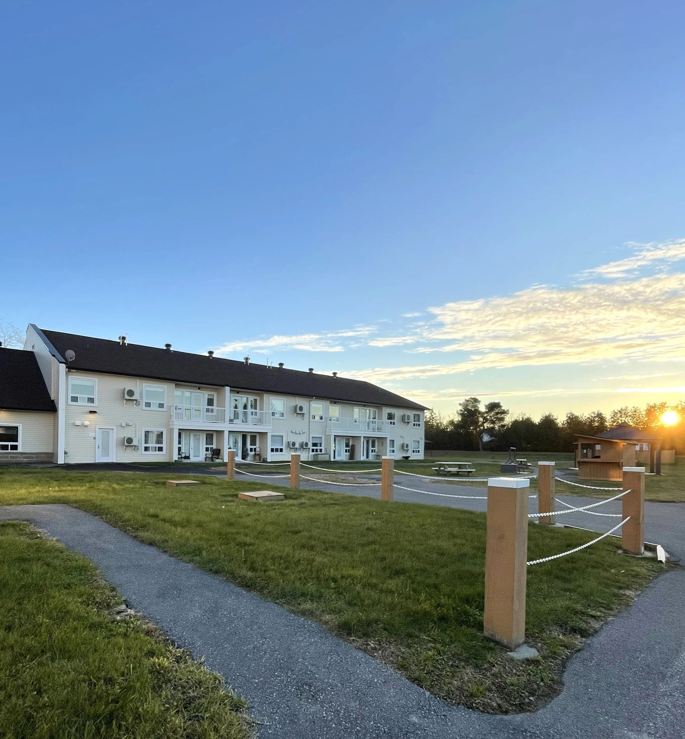 A two-story apartment building with balconies, surrounded by a grassy area with sidewalk paths, a small parking lot, and a sun setting in the distance.