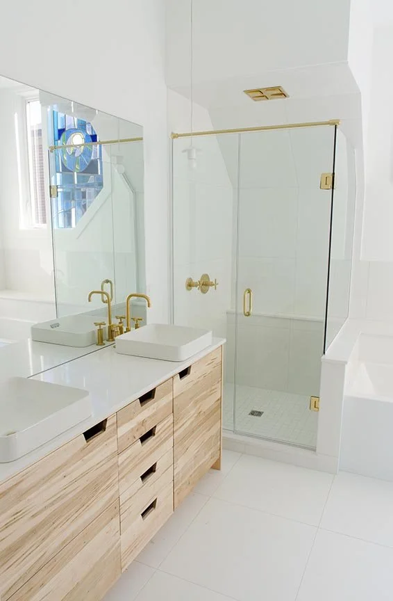Modern bathroom with a wooden vanity, white countertops, and gold fixtures. Glass shower enclosure with gold hardware and a white tile interior. Large mirror above the vanity and a window providing natural light.