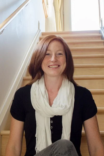 A woman with red hair smiles while sitting on wooden stairs inside an office with a large window and cream-colored curtains.