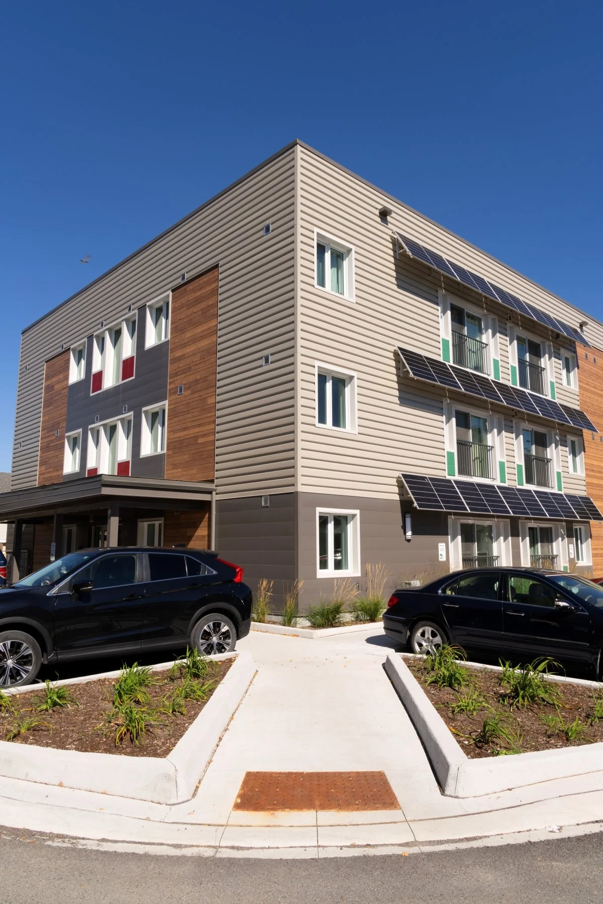 an image of a 3 storey affordable apartment building with metal siding, solar panel shade awnings on the south elevation, concrete walkways with landscaped planters separating parking from concrete sidewalks, a deep covered porch at the main entry
