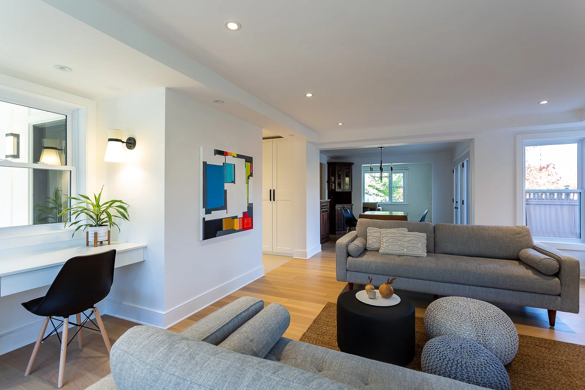 Open-concept living room with gray sofas, modern artwork, a window seat with a black chair and a window, wooden flooring, and a dining area visible in the background.