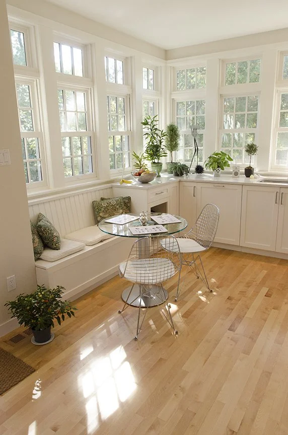 Bright kitchen nook with white cabinetry, multiple large windows, a built-in window seat with cushions, a glass round table, metal wire chairs, and potted plants on the windowsill.