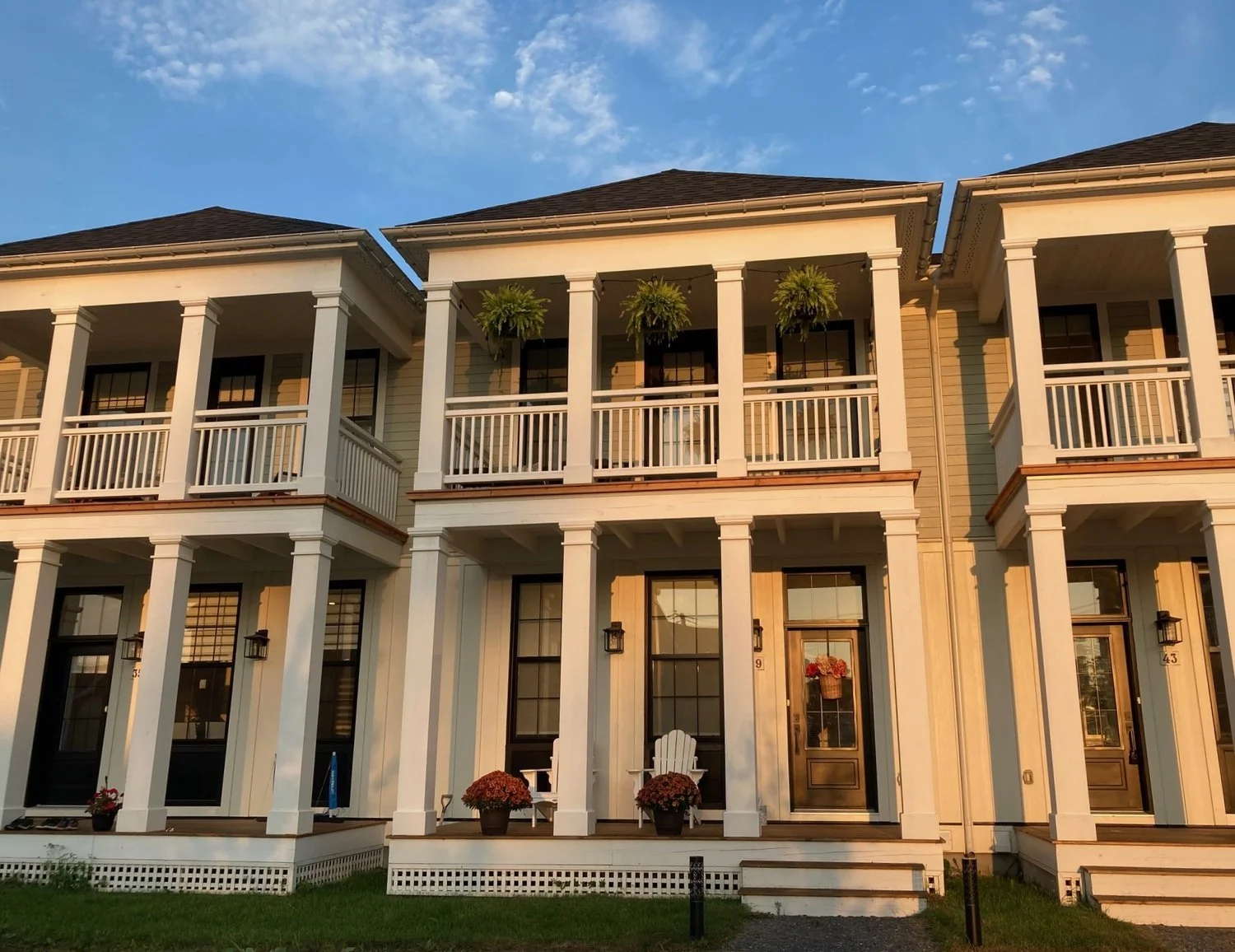 Three-story row of modern townhouses with balconies, white railings, potted plants, and a porch with two white chairs, under a blue sky.