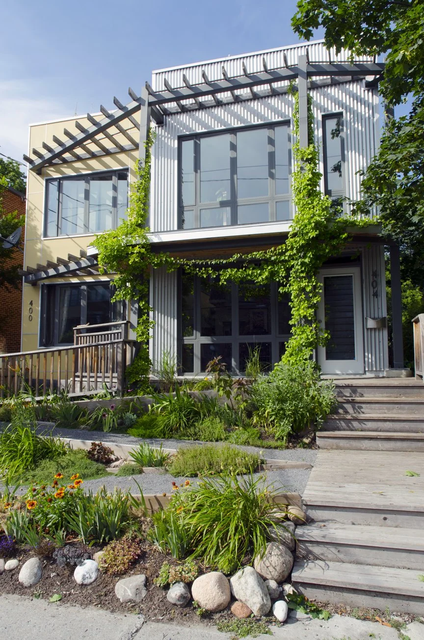 Modern two-story mixed use residential and commercial building with large glass windows, metal siding, and greenery climbing the exterior, surrounded by a landscaped garden with accessible ramp, and a wooden staircase leading to the entrance.