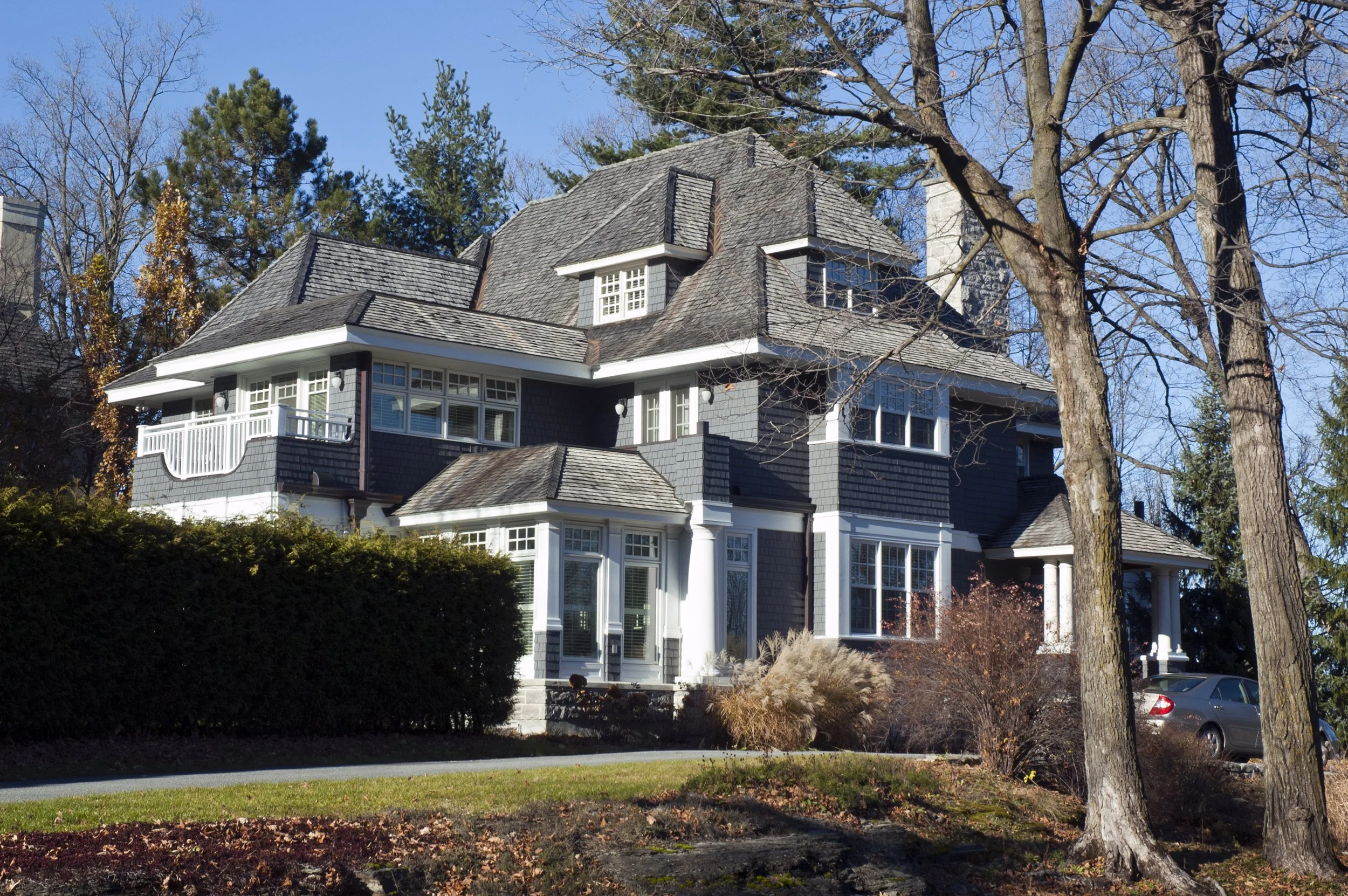 Large, multi-story house with dark gray exterior, multiple windows, and a sloped roof, surrounded by trees and landscaping.
