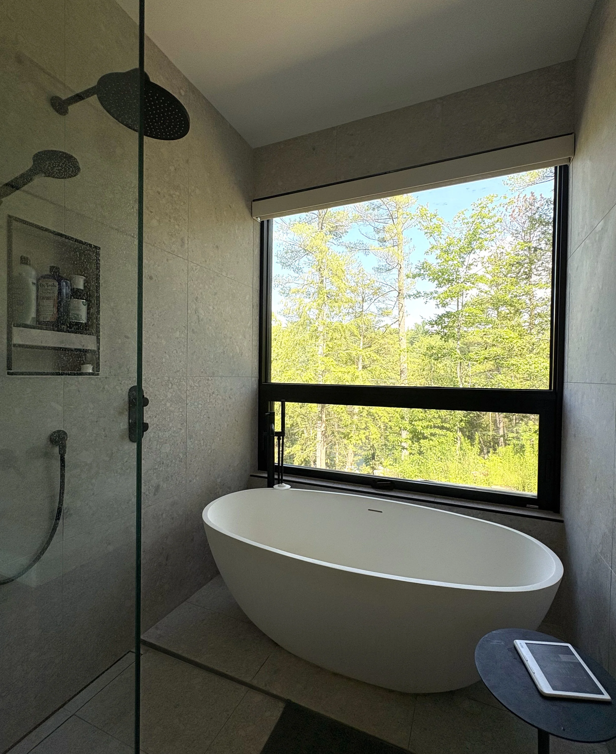 Modern bathroom with a freestanding bathtub near a large window overlooking a forest, with a shower area on the left and a small round table with a tablet on it.