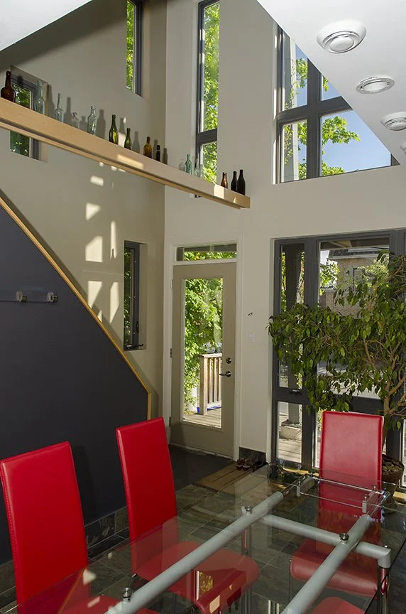 Modern office meeting area with three red chairs around a glass table, large windows, a door leading outside, and decorative bottles on a shelf above.