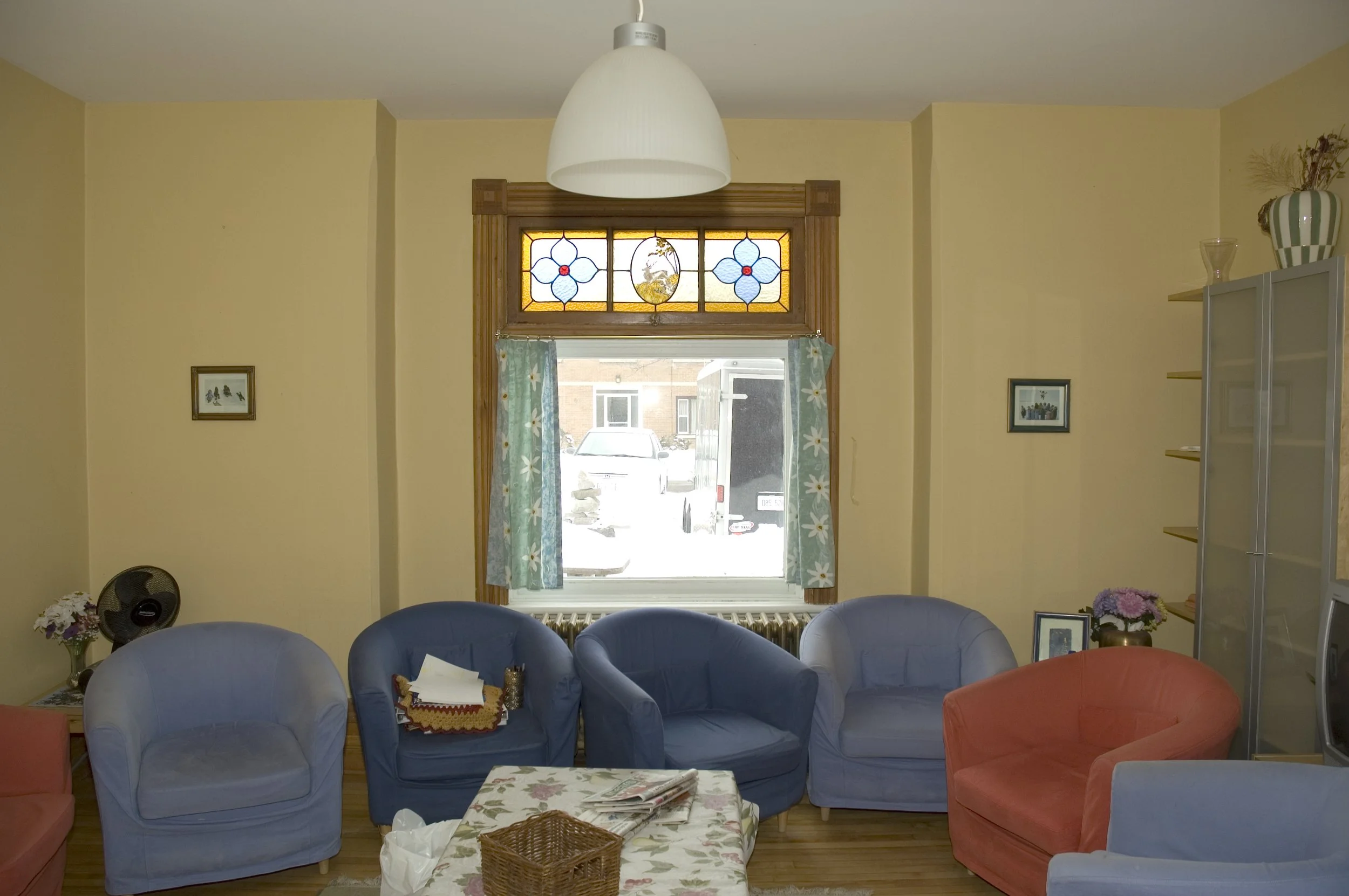 Living room with yellow walls, colorful armchairs, a window with floral curtains, and stained glass above the window.