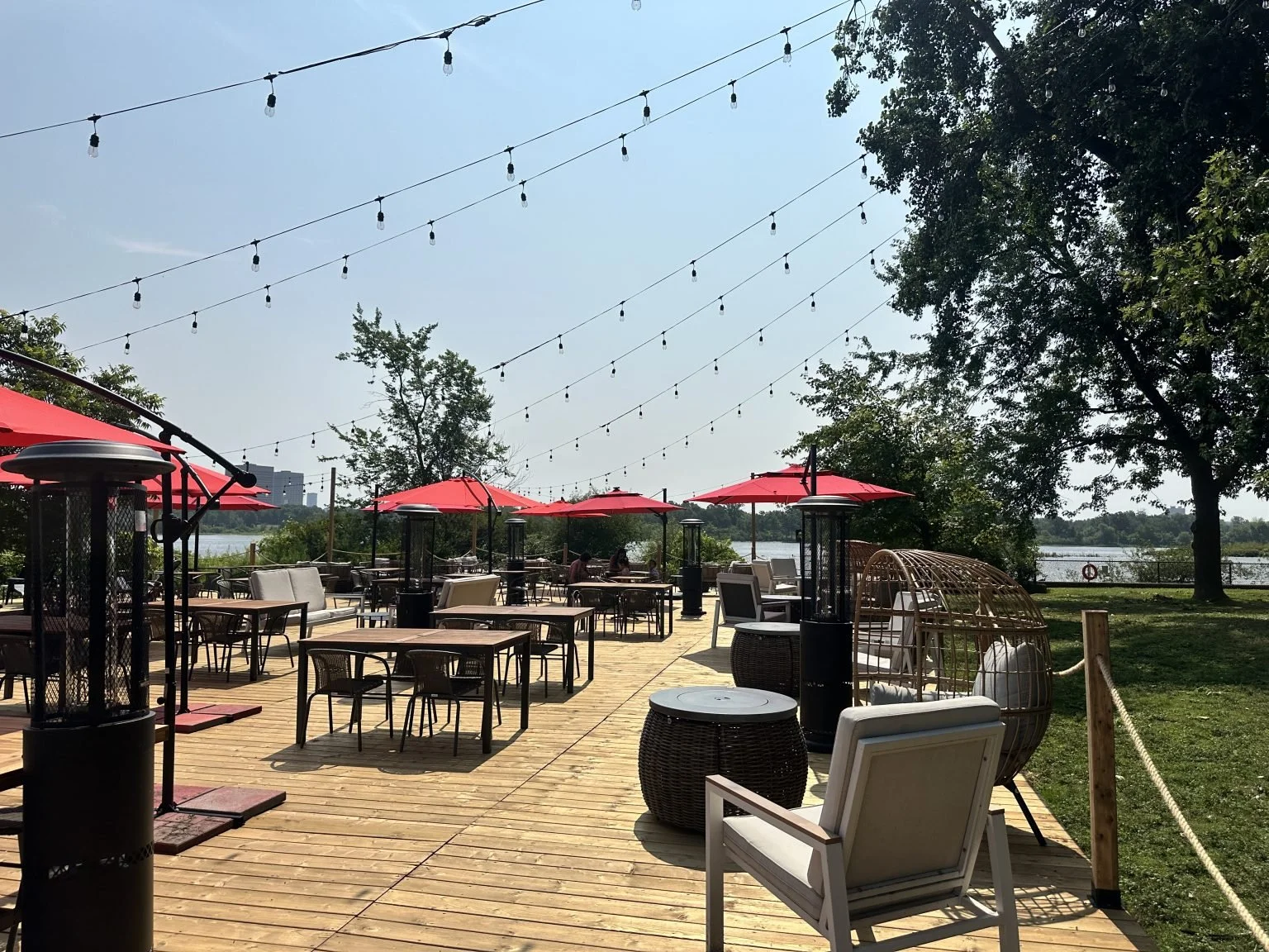 Outdoor patio with tables, chairs, and red umbrellas near a body of water, decorated with string lights, with trees and a clear sky in the background.