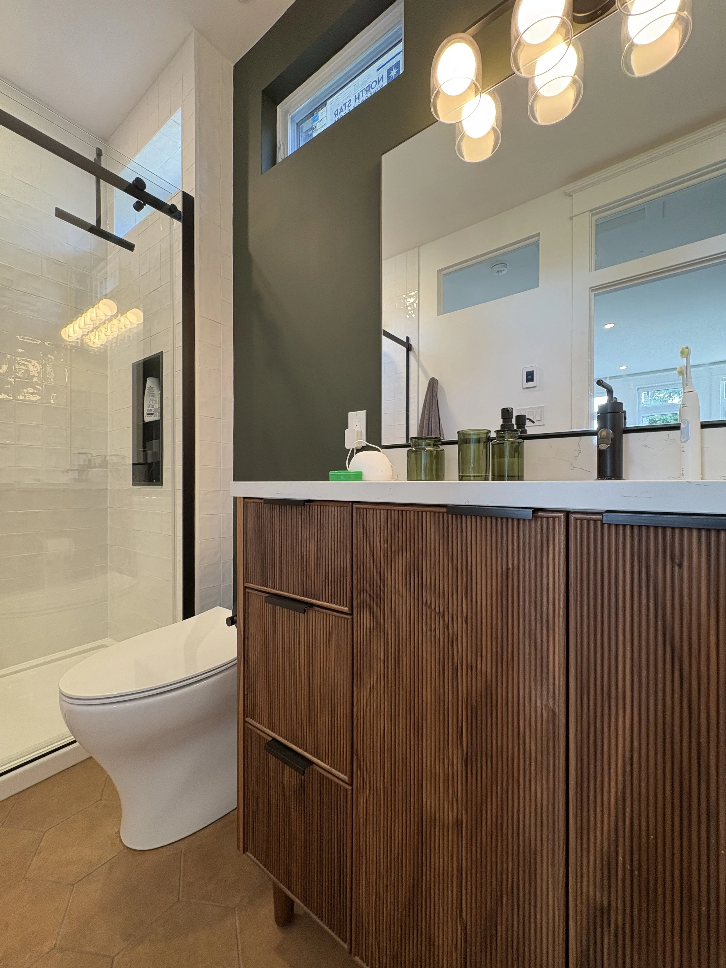 A modern bathroom with a wooden vanity, a large mirror, and a walk-in shower with glass doors. The vanity has dark hardware, and the countertop holds various toiletries in green containers and a soap dispenser. Recessed and ceiling lights illuminate the room.