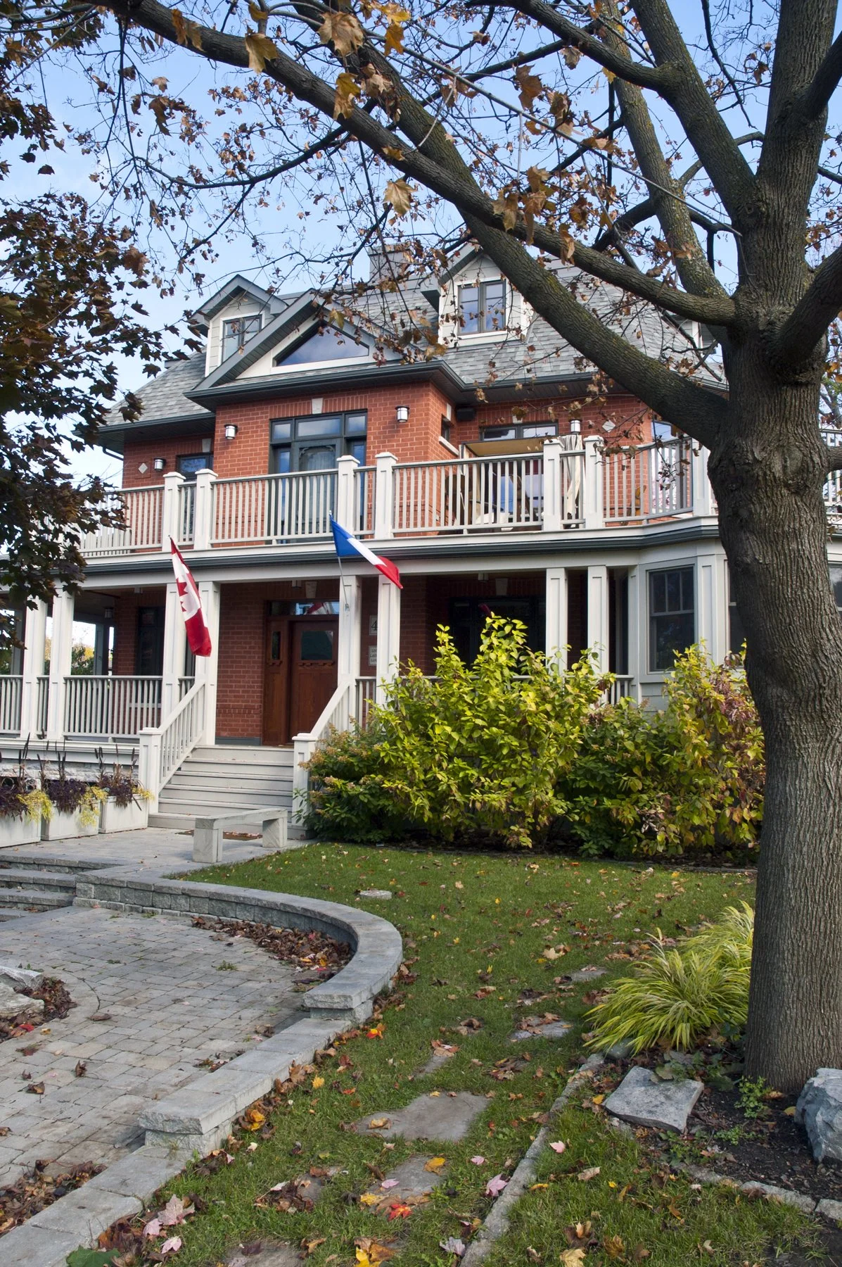A multi-story brick house with balconies, flags, a small front porch, and a landscaped yard with trees and fallen leaves.