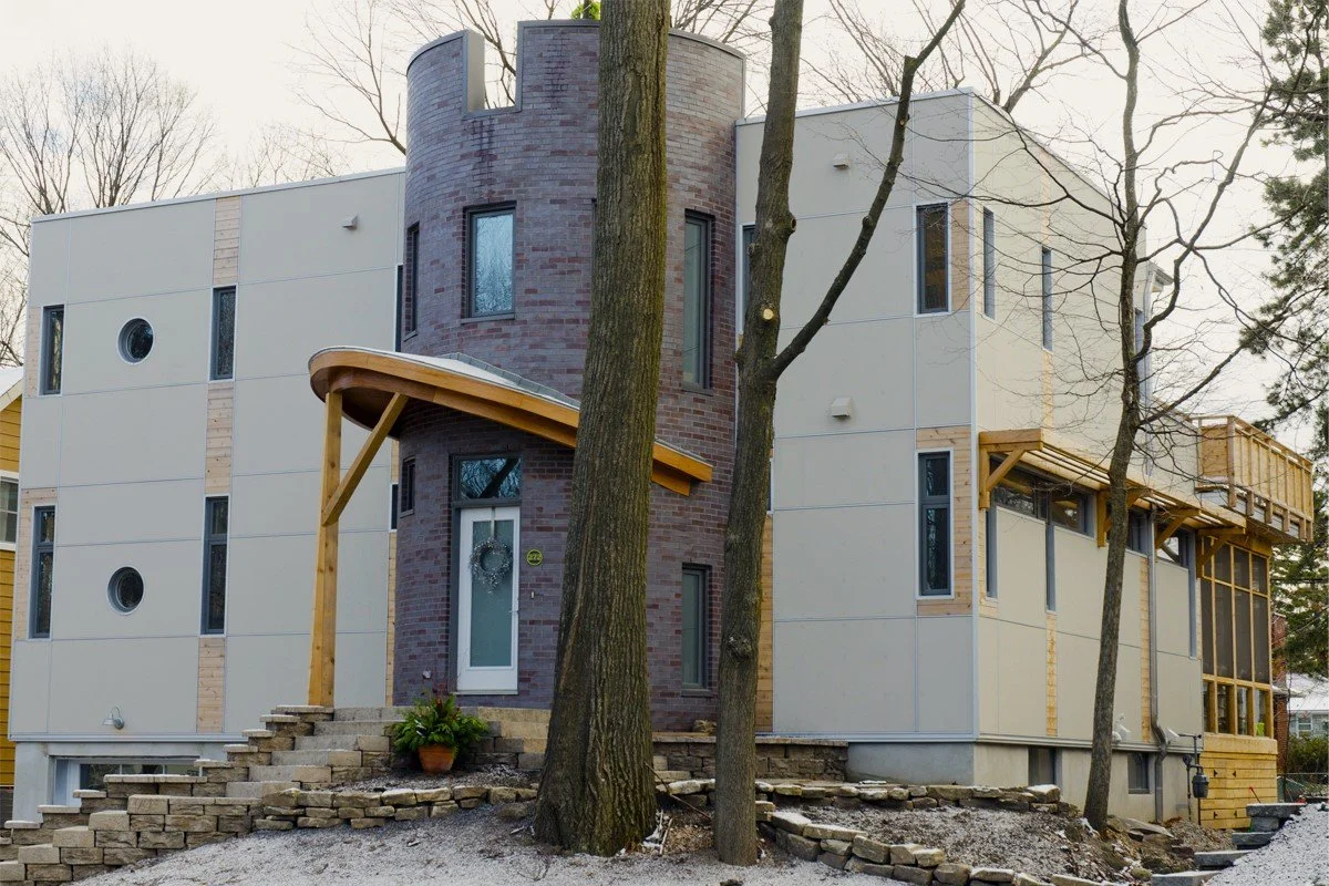Modern home under construction with a mix of brick and siding exterior, wooden accents, and vertical windows, surrounded by trees and stone landscaping.