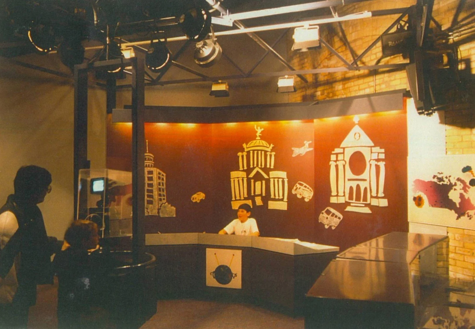 People standing in front of a news studio with a news anchor seated at the desk against a backdrop of cityscapes and historical landmarks.