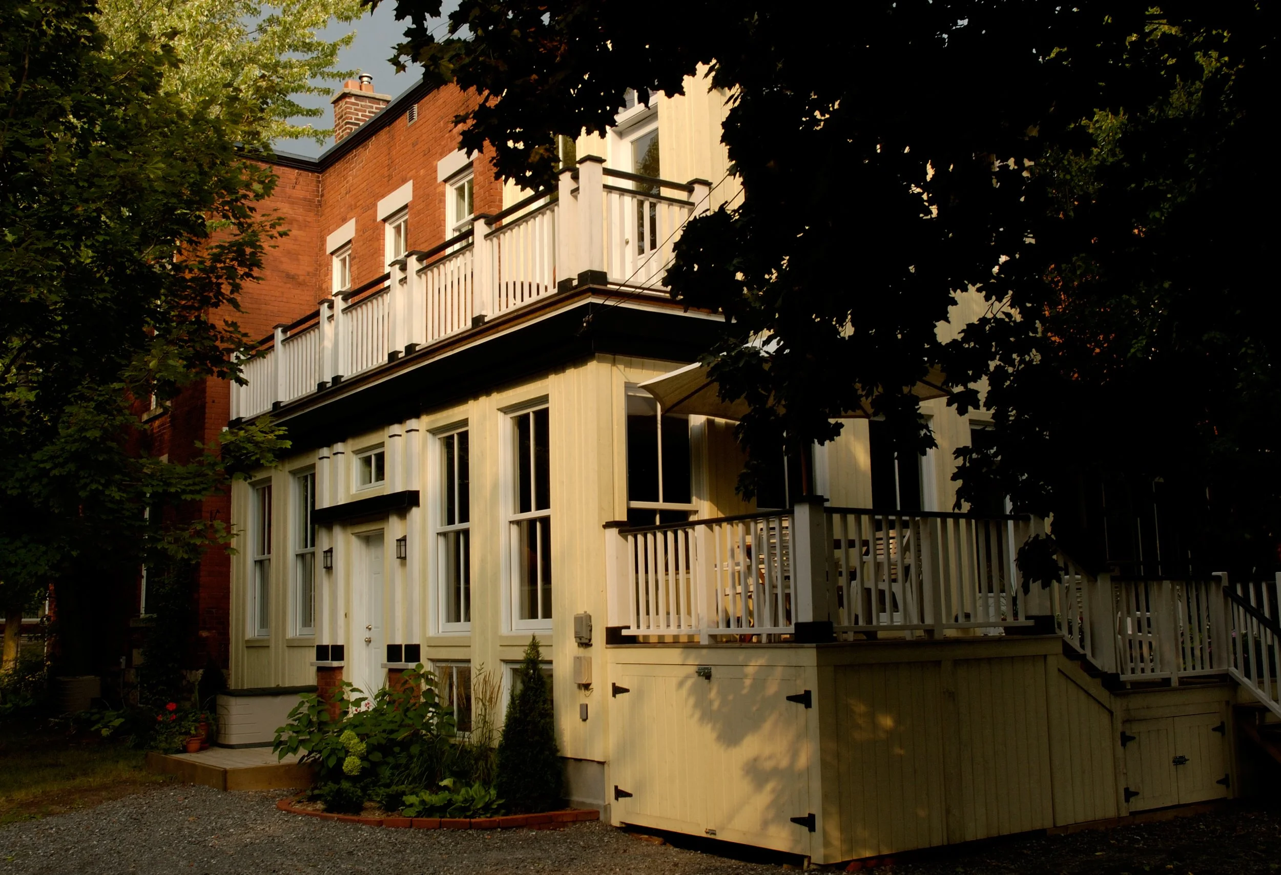 A multi-story house with beige wooden siding, multiple large windows, and a white balcony with black accents, surrounded by trees.