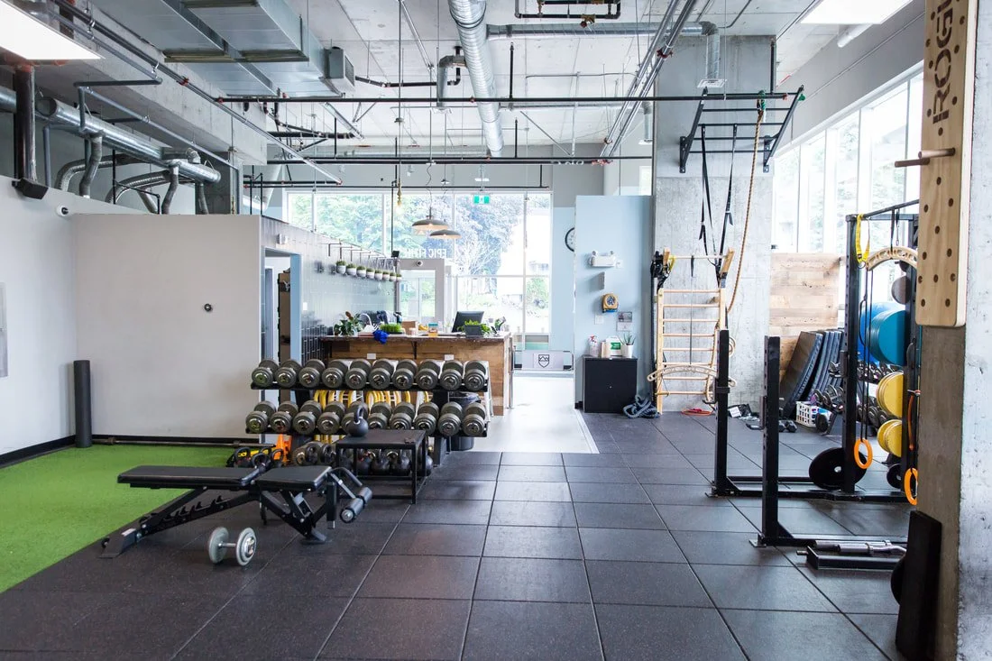 Commercial gym with weights, benches, resistance bands, and workout equipment, with large windows and industrial ceiling.